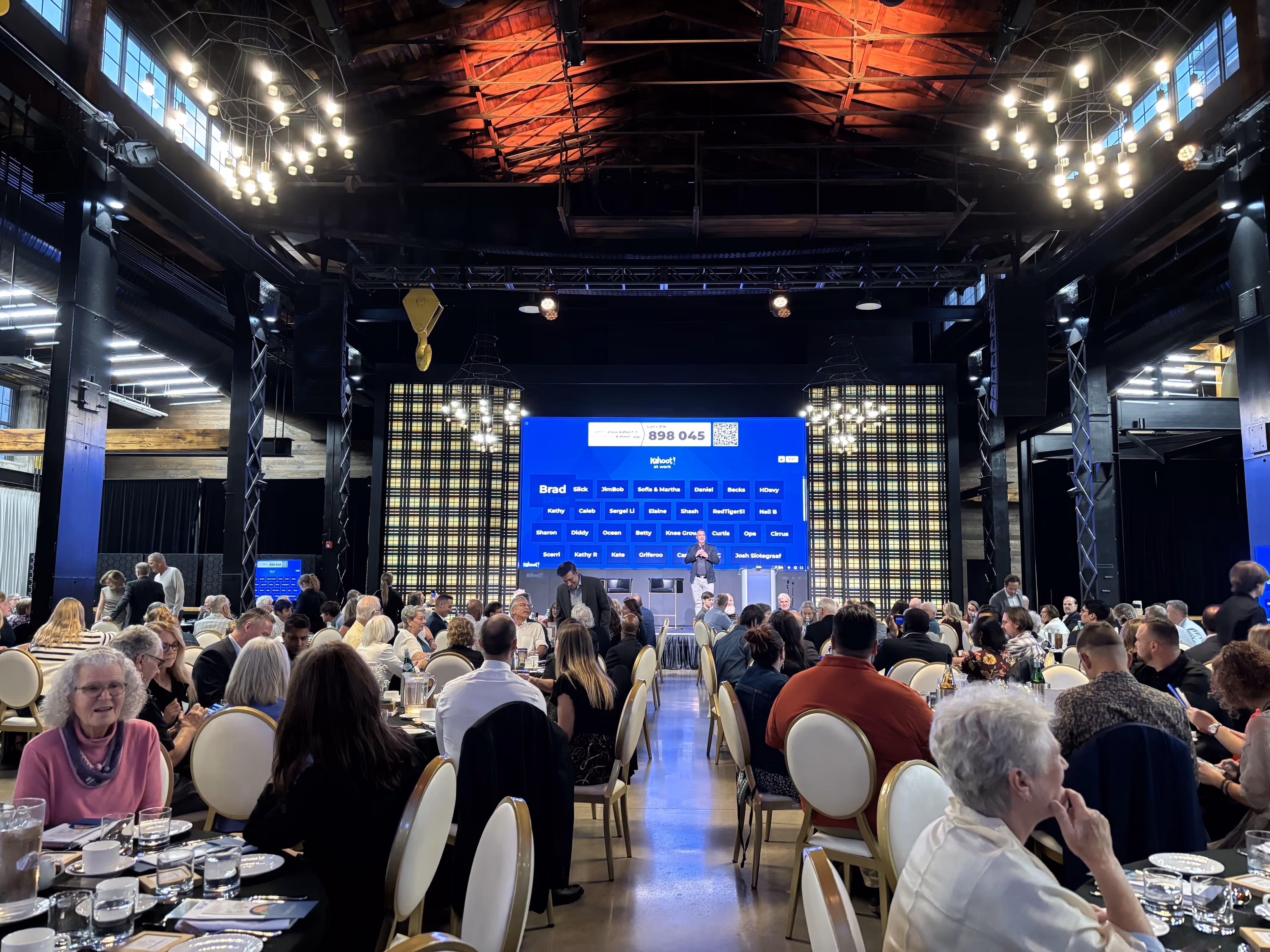 Large group of people seated at round tables in a banquet hall, with a presenter speaking in front of a large screen displaying names and scores.