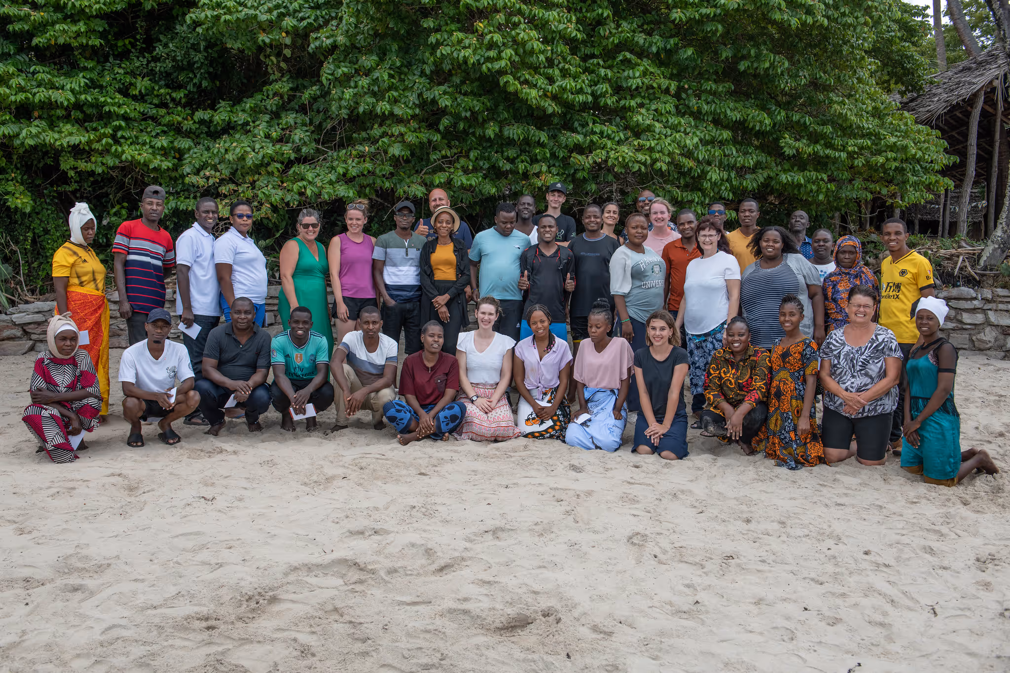 Group photo of diverse people standing and sitting on sandy ground with dense green foliage in the background.