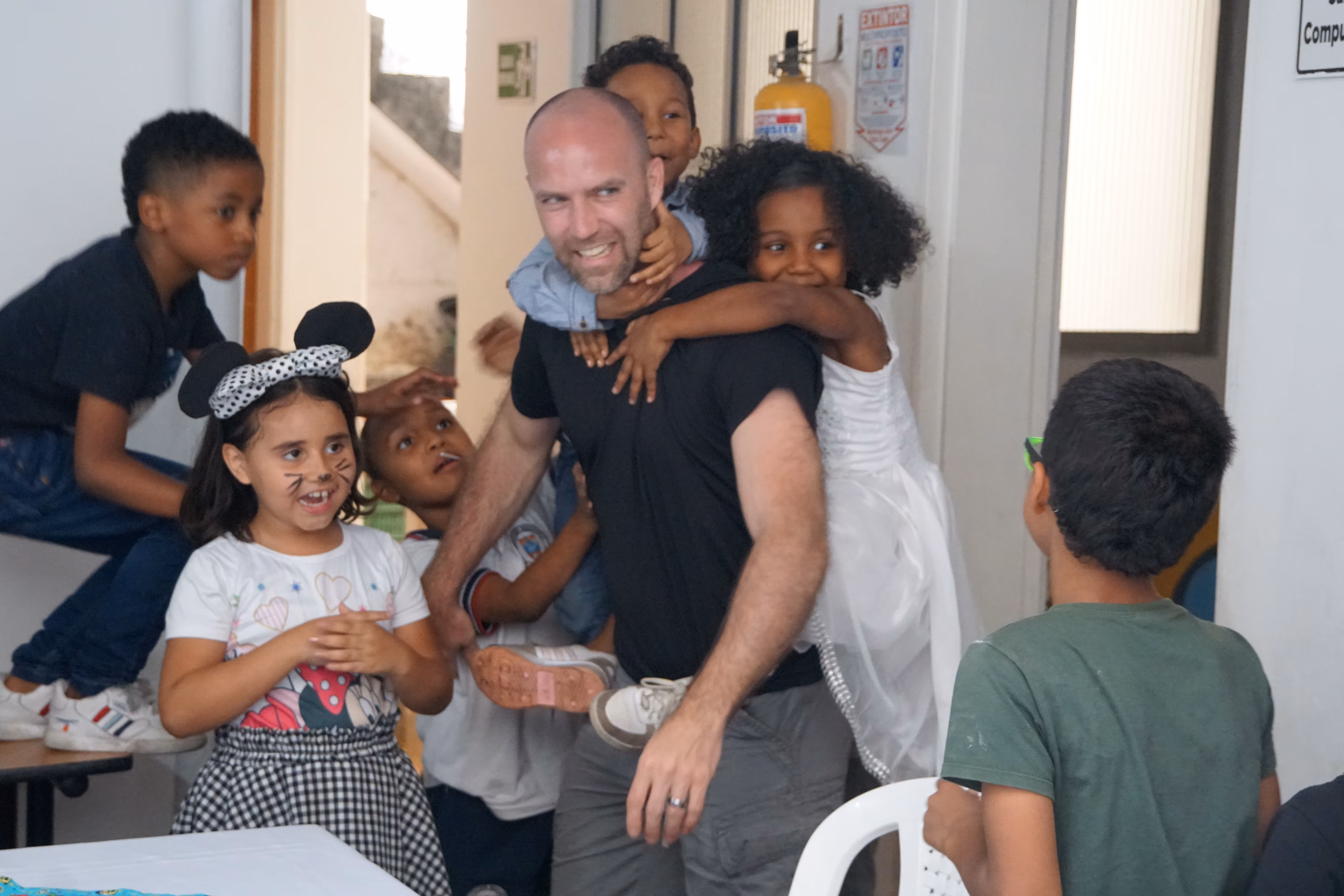 Smiling man carrying a girl in a white dress on his back surrounded by happy children, one dressed with mouse ears and face paint.