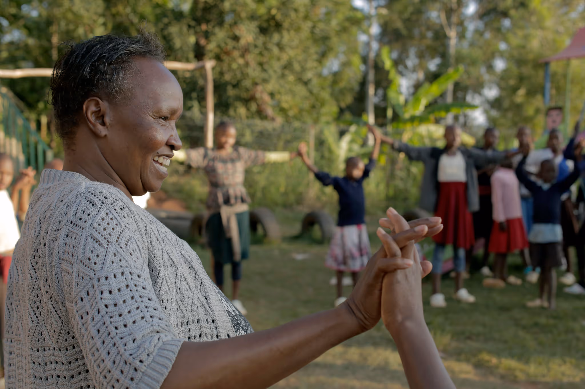 Smiling woman holding hands with a child outdoors with a group of children holding hands in the background.