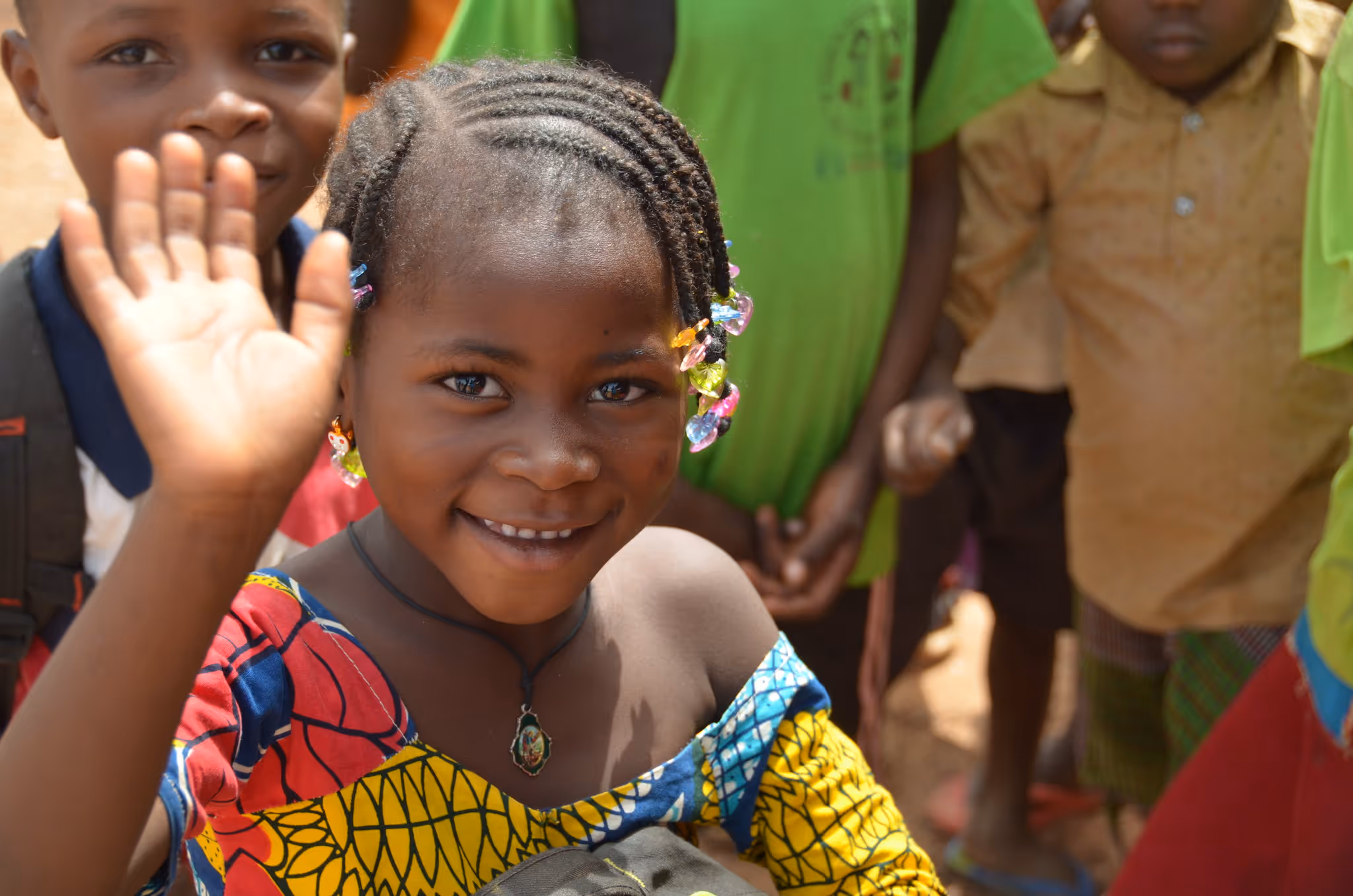Smiling young girl with colorful beaded hair waves at the camera, surrounded by other children.