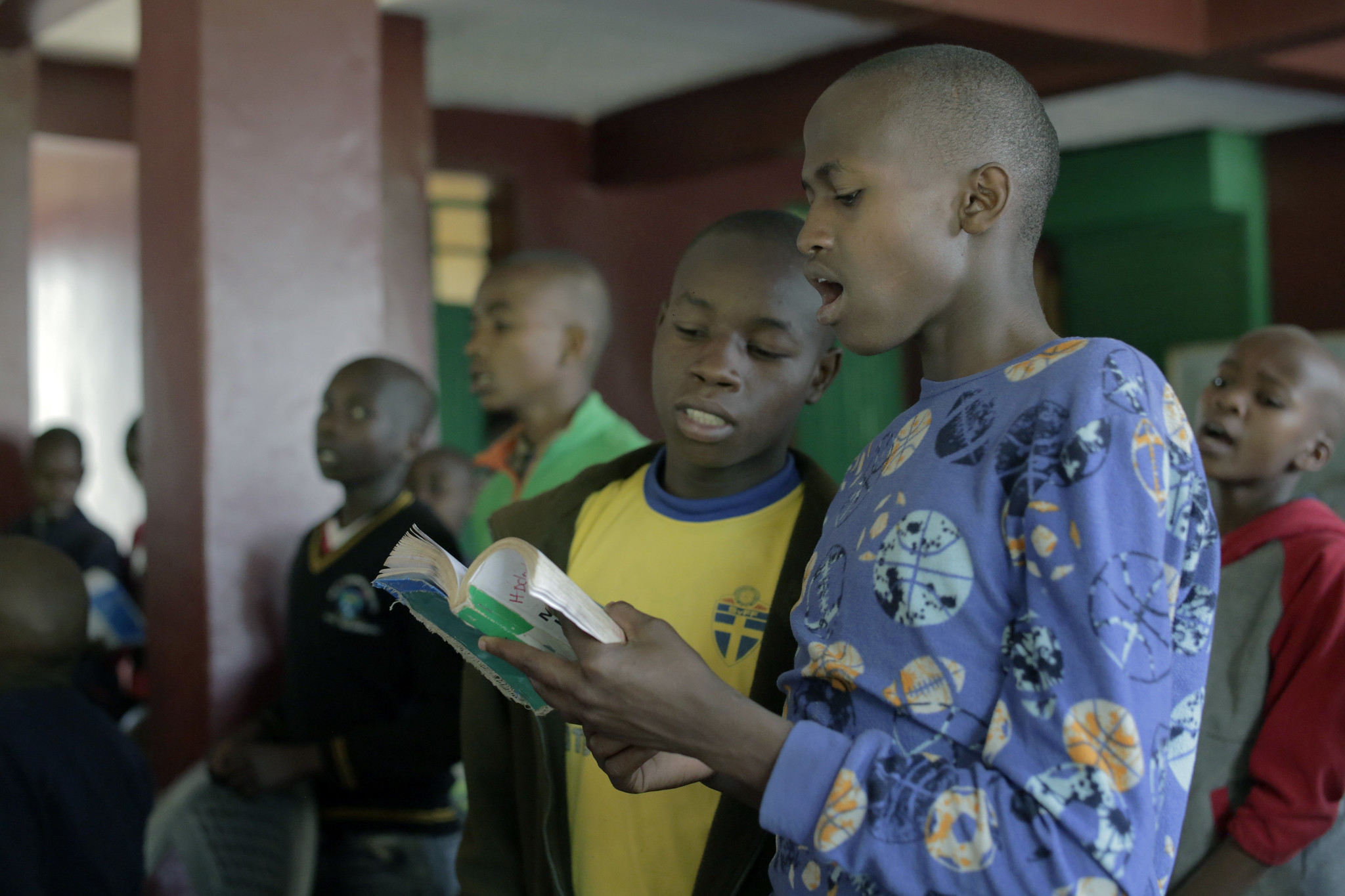 Two boys reading aloud from a book while others listen attentively in a classroom setting.