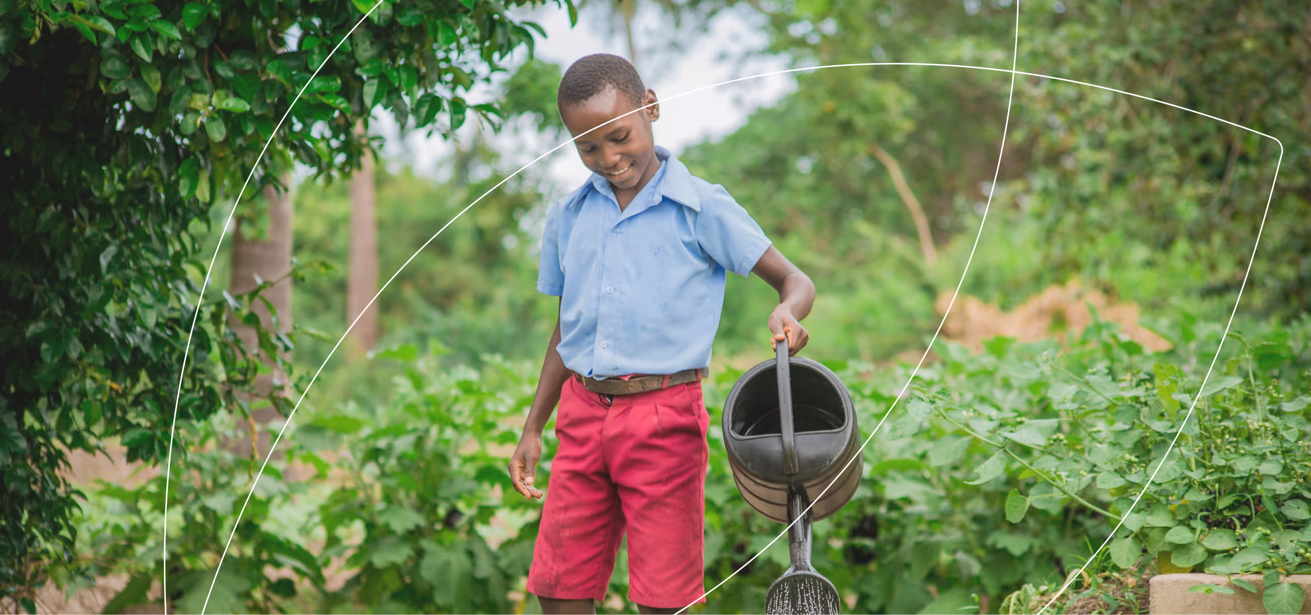 Boy in blue shirt and red shorts watering plants in a garden with a black watering can.