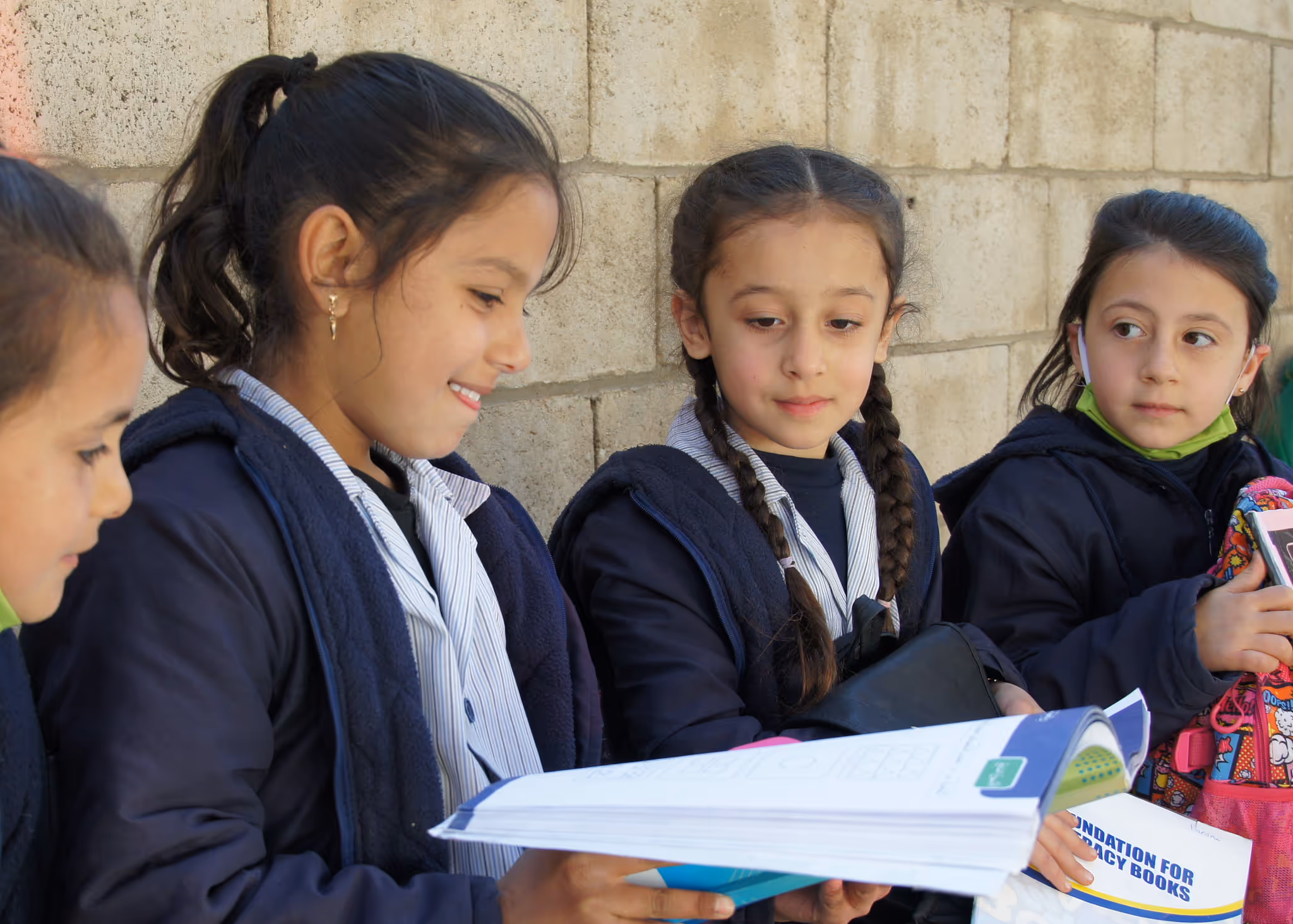 Four children sitting indoors on stools and the floor, reading books against a wall painted with trees and a sun.