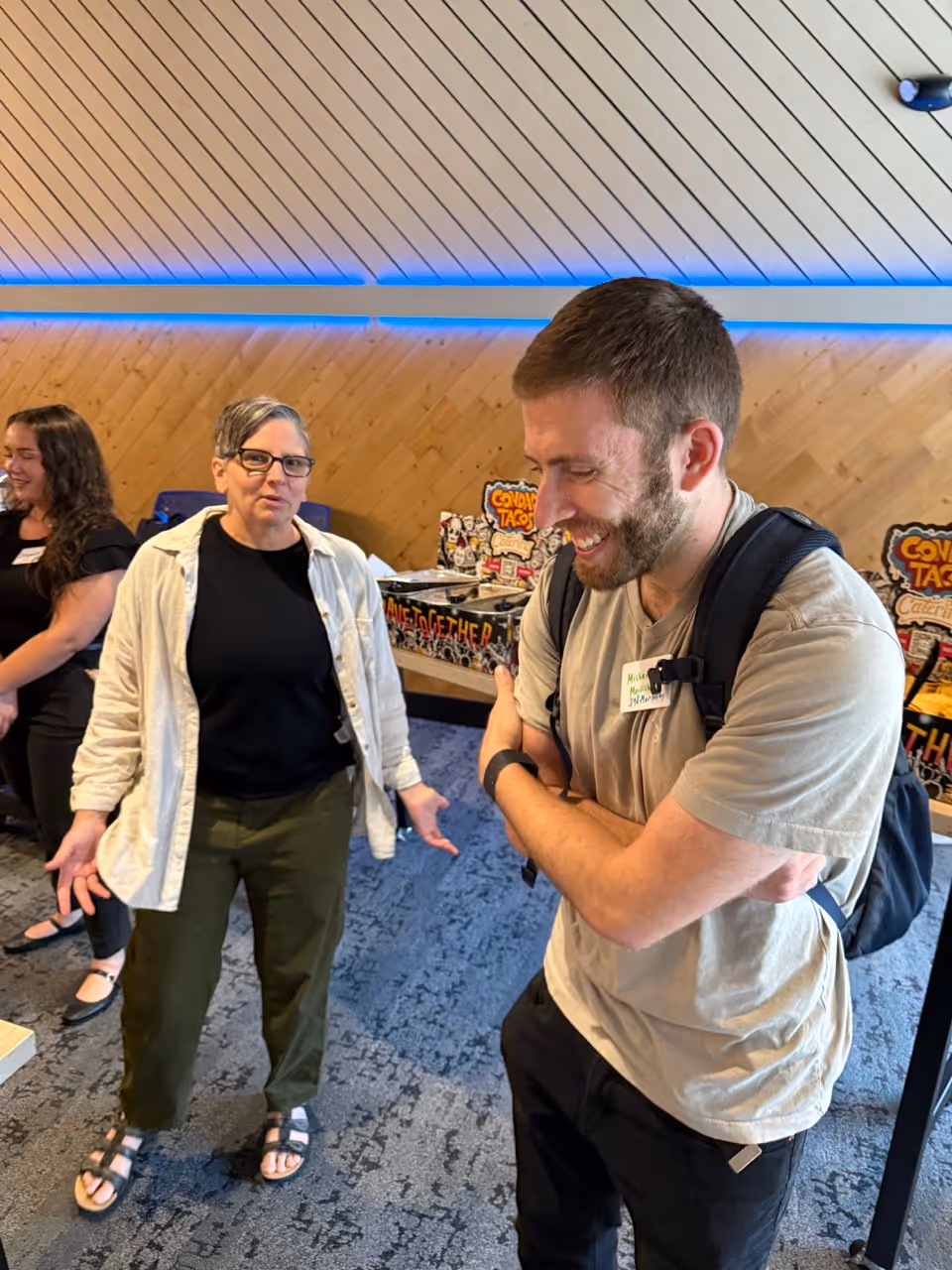 Three people casually interacting indoors; a woman with short gray hair and glasses gestures while a bearded man smiles with arms crossed, and another woman stands in the background.