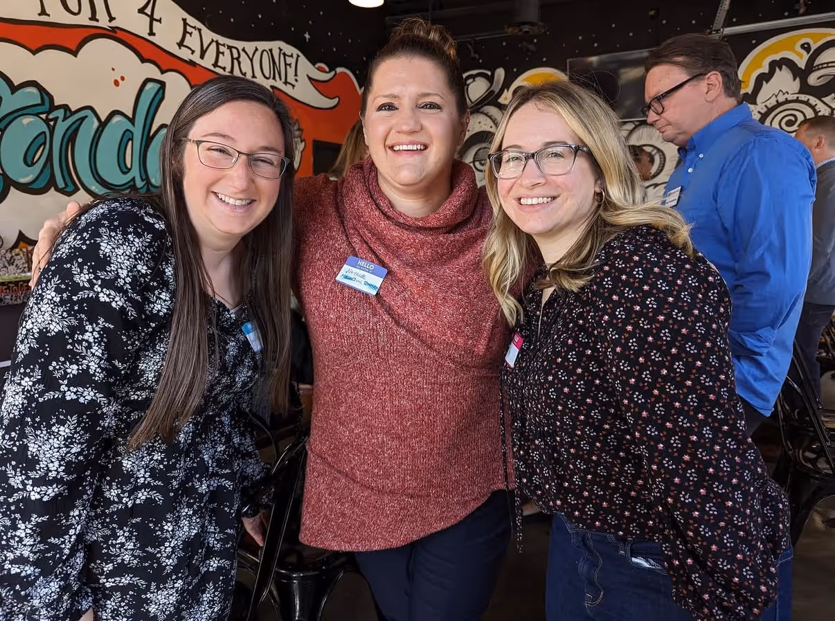 Three smiling women wearing glasses, standing closely together indoors with colorful graffiti on the wall behind them.