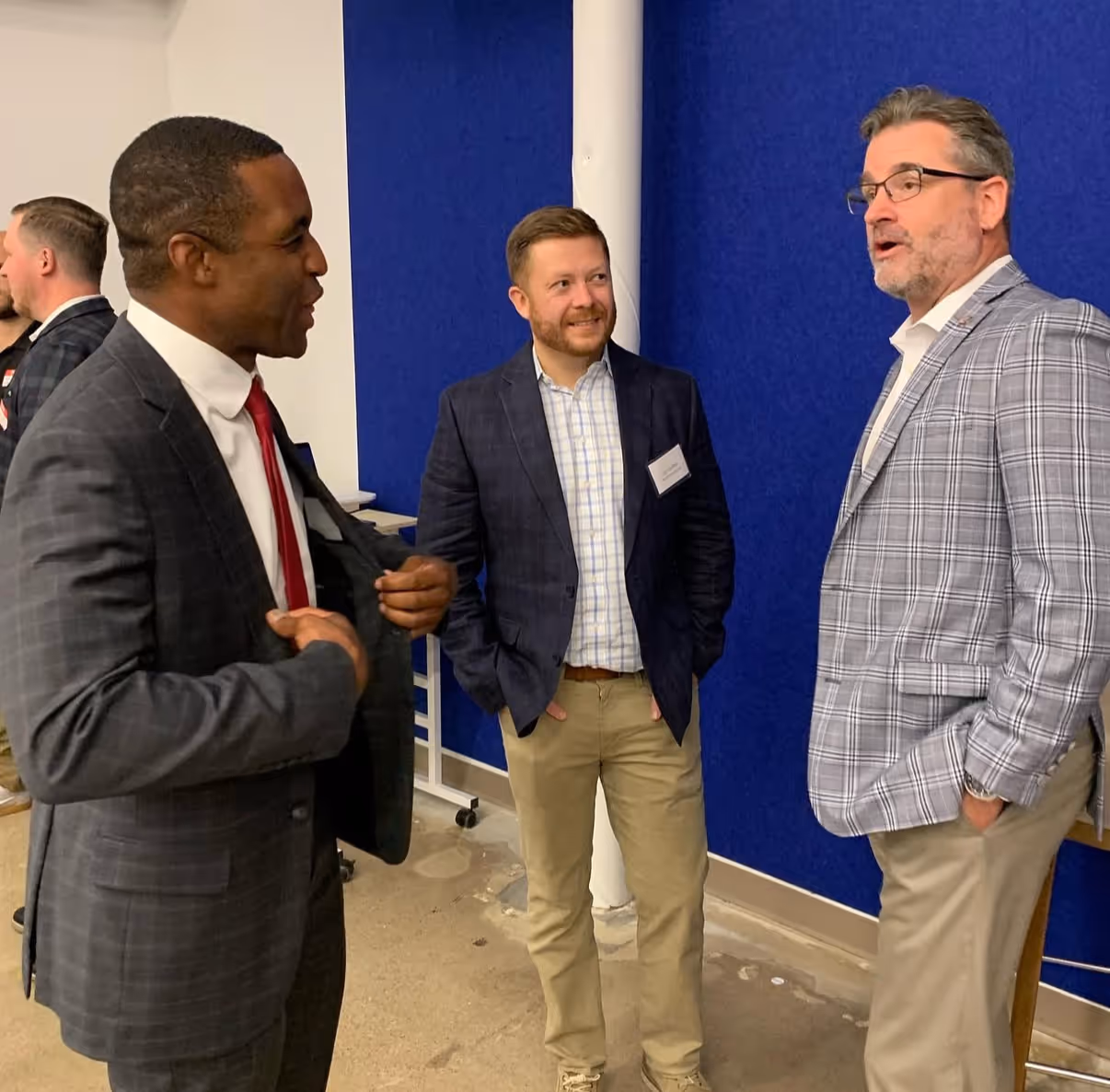 Three men in business casual attire having a conversation in front of a blue wall.