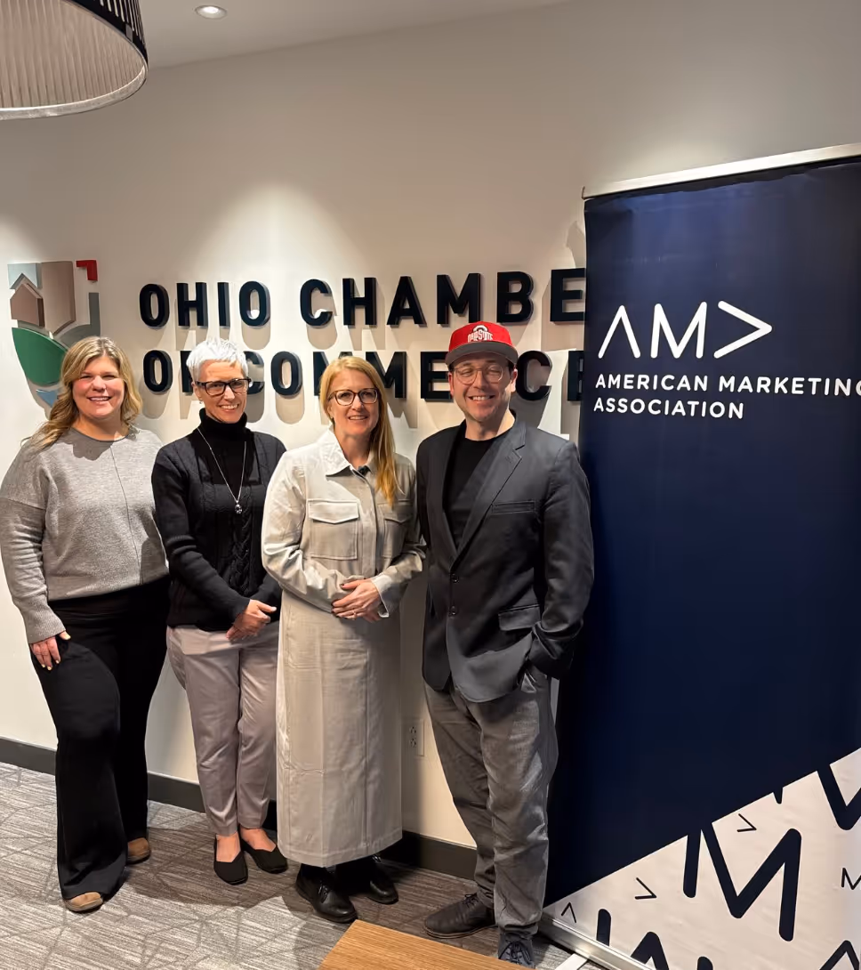 Four people standing indoors in front of an Ohio Chamber of Commerce sign, next to a blue banner for the American Marketing Association.