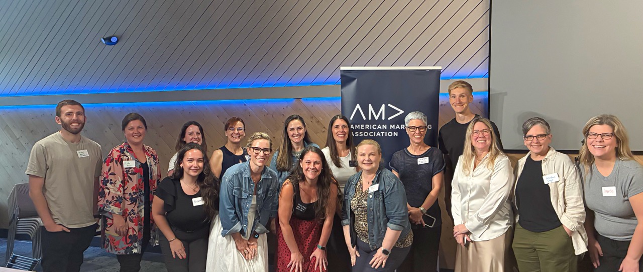 Group photo of 15 smiling adults standing indoors in front of an American Marketing Association banner.
