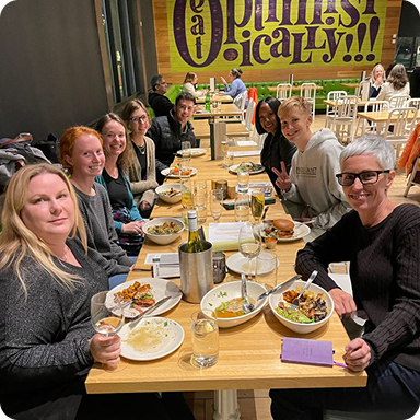 Group of eight people sitting around a rectangular wooden table with plates of food and drinks in a casual restaurant.