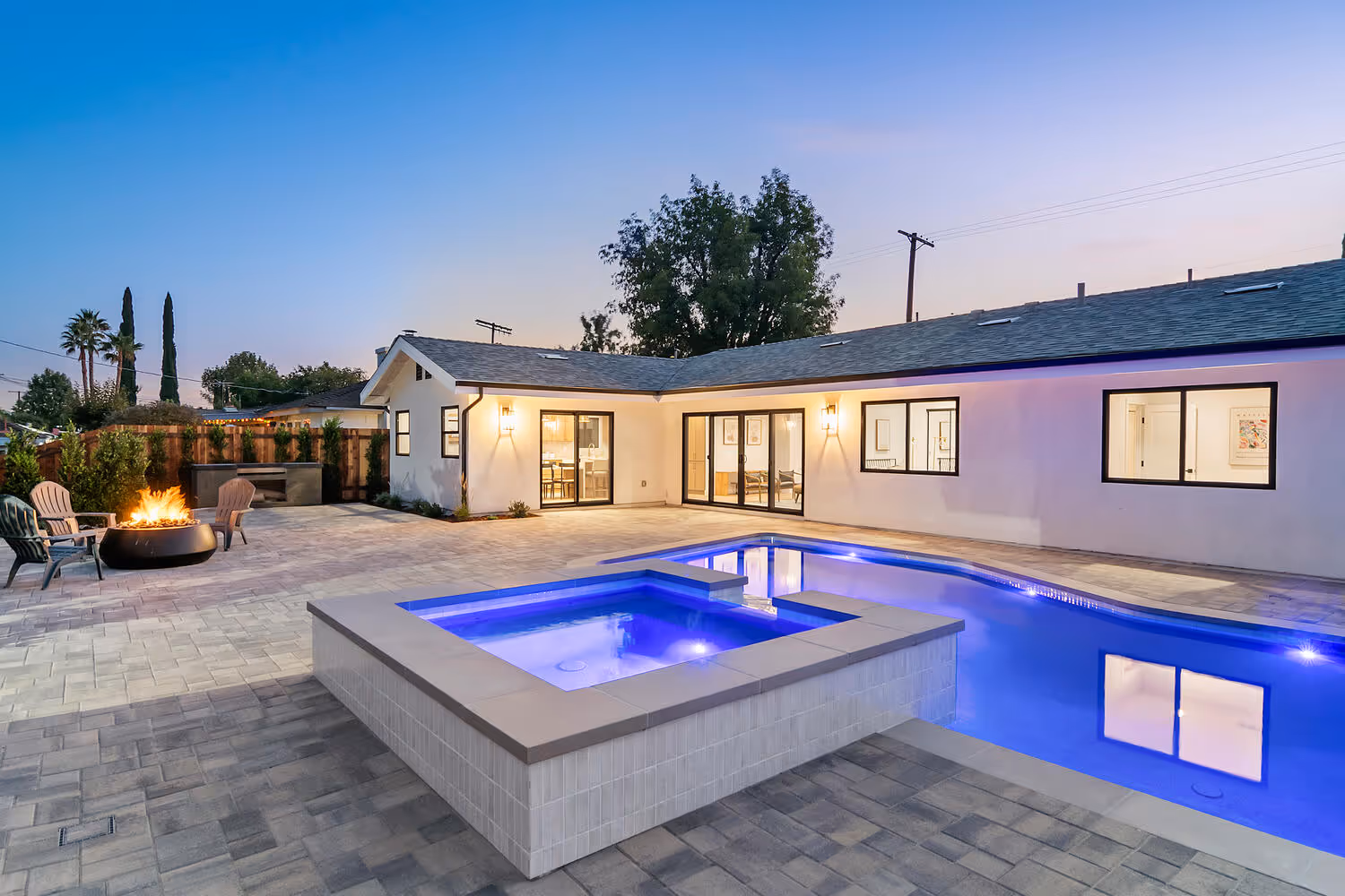 Modern backyard with illuminated pool, spa, and fire pit at twilight