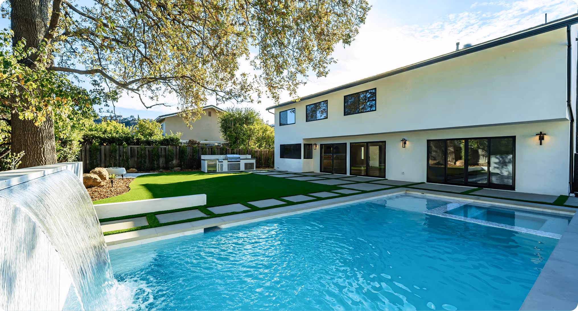 Modern backyard with swimming pool, waterfall, and outdoor kitchen under tree
