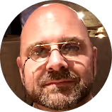 Bald man with glasses and beard smiling warmly in circular portrait