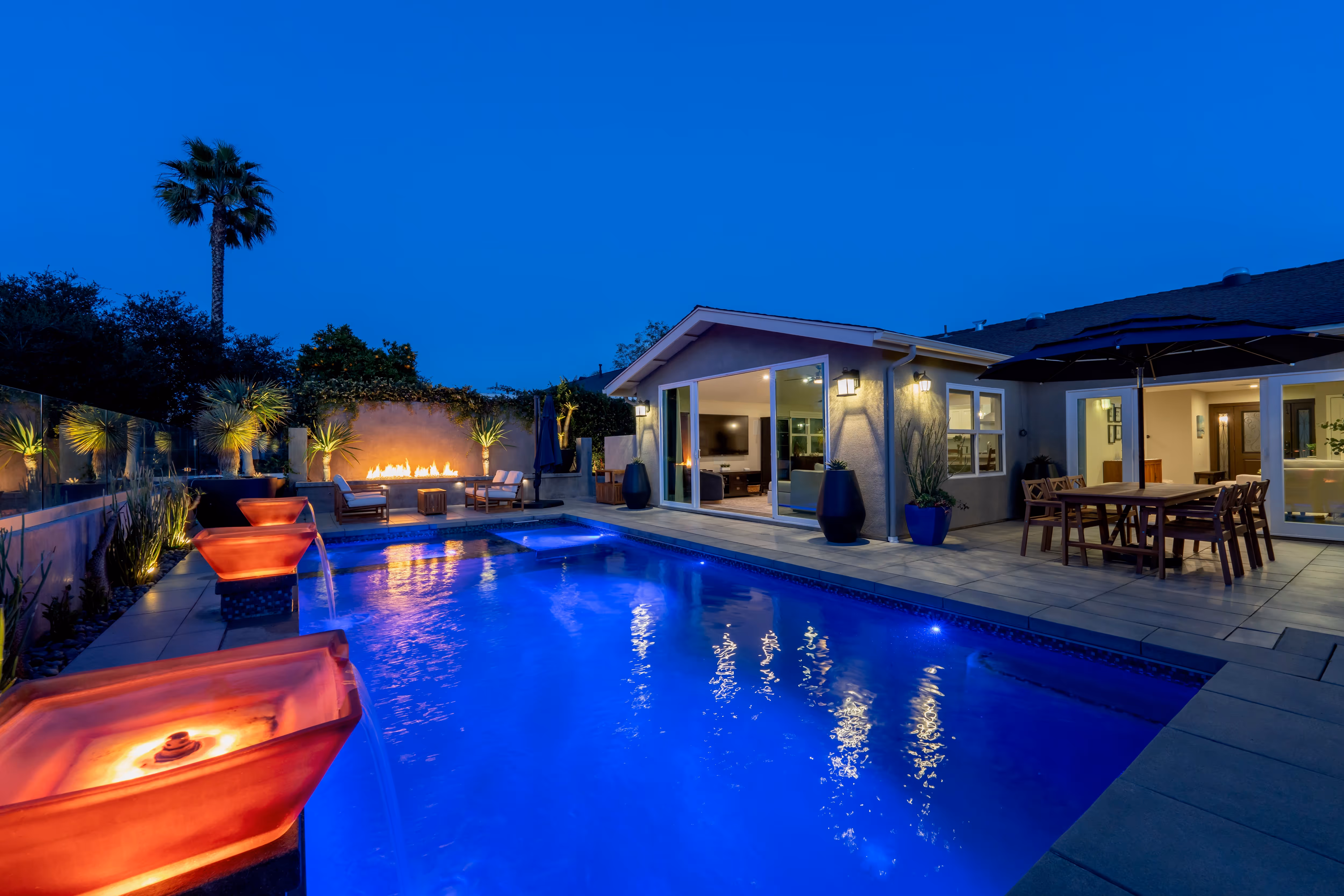 Luxurious backyard with blue swimming pool, fire features, and palm tree at night