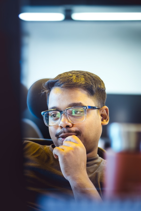A focused young man wearing glasses works intently at his computer in a modern office, bathed in soft blue lighting that highlights a thoughtful and tech-savvy atmosphere.