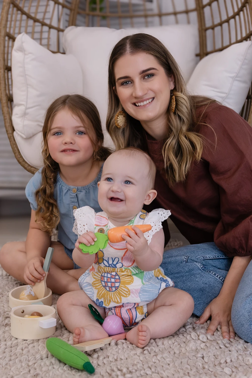 A happy woman sits with two young children, one playing with toy food. They are surrounded by soft cushions on a light rug.