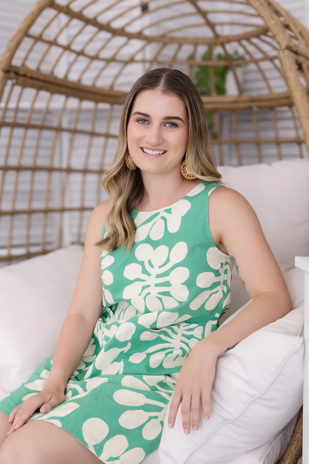 Smiling woman with long hair wearing a green and white patterned sleeveless dress sitting on a white cushioned chair with a wicker backrest.
