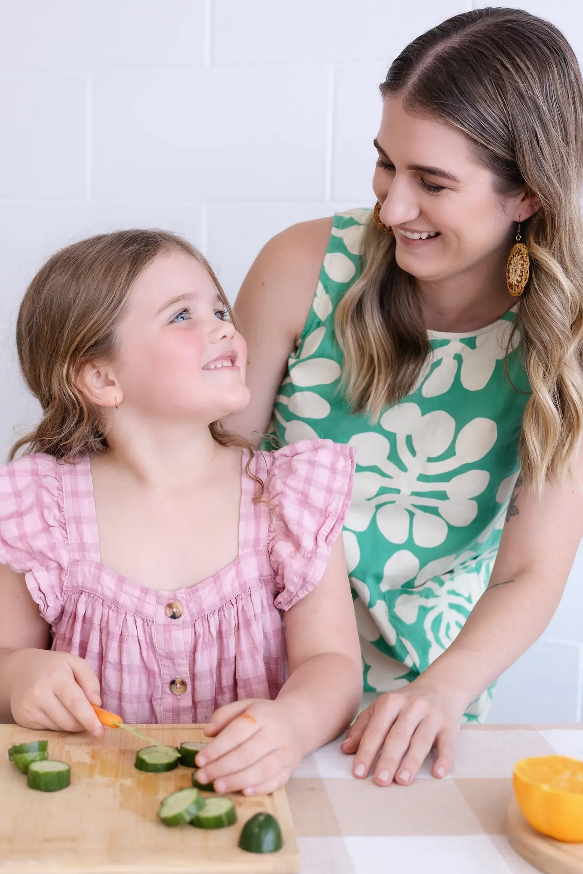 Young girl smiling at a woman while cutting cucumber slices on a wooden board in a kitchen.
