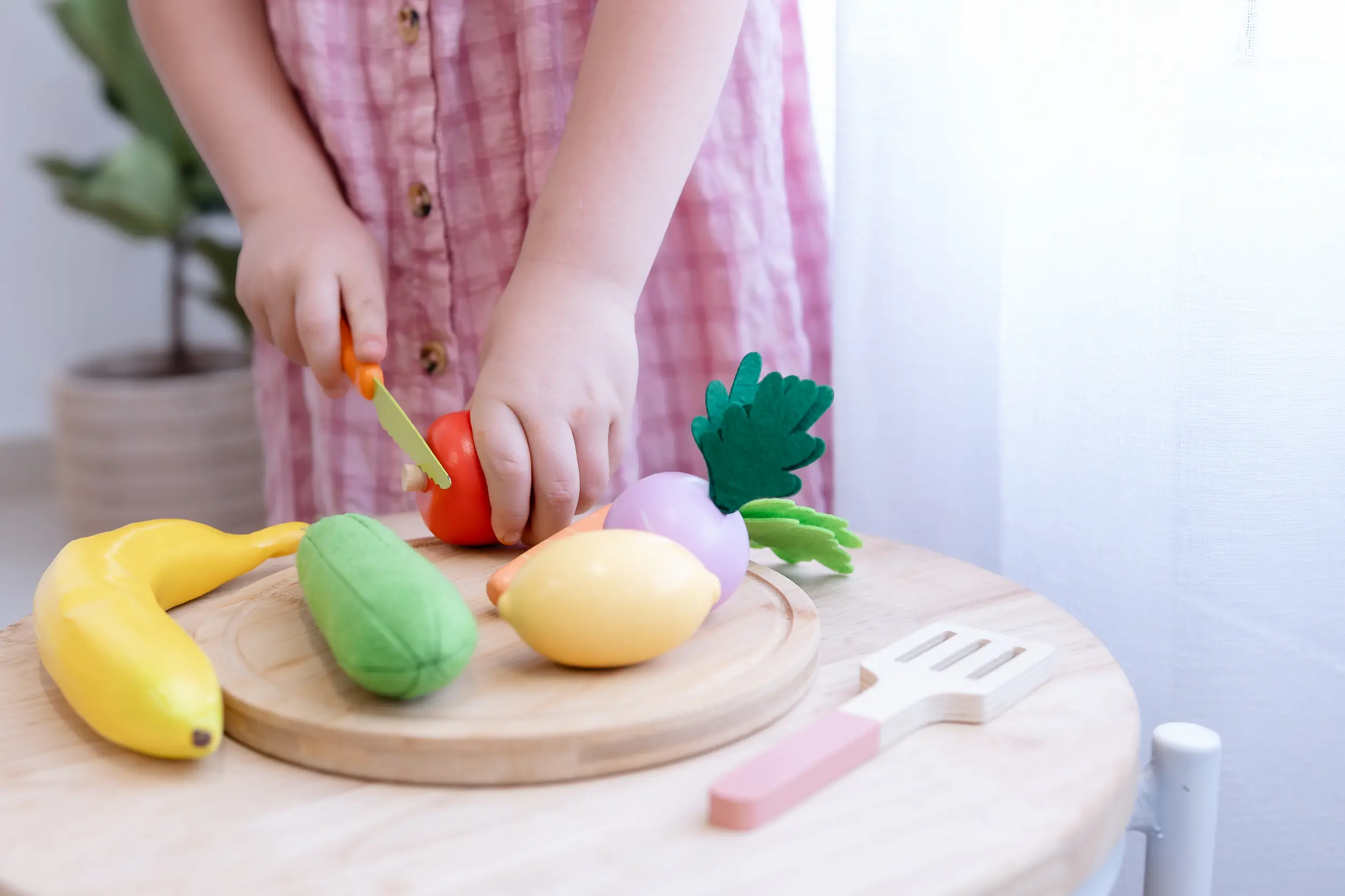 Child in a pink dress cutting a toy tomato with a toy knife on a wooden board with other toy fruits and vegetables nearby.