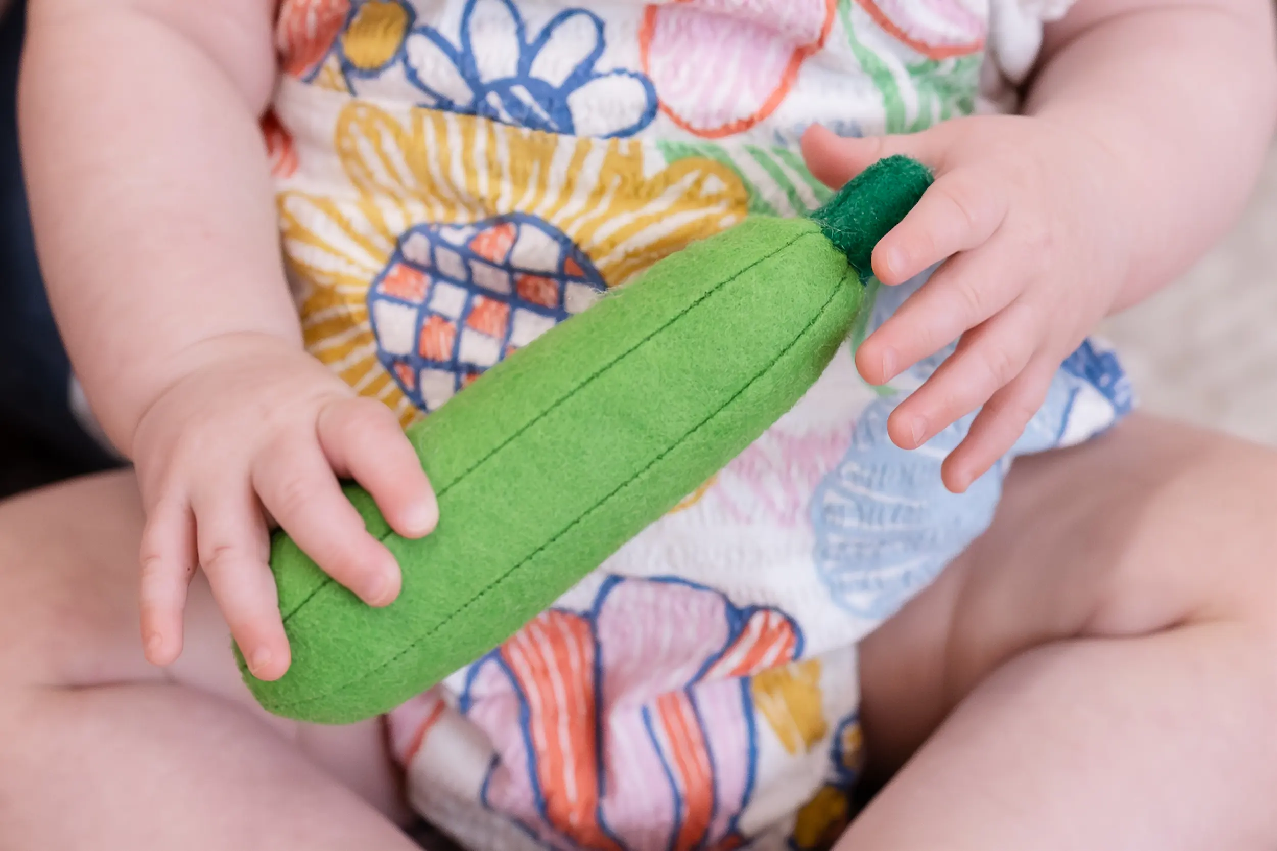Baby wearing colorful floral outfit holding a soft green toy cucumber.