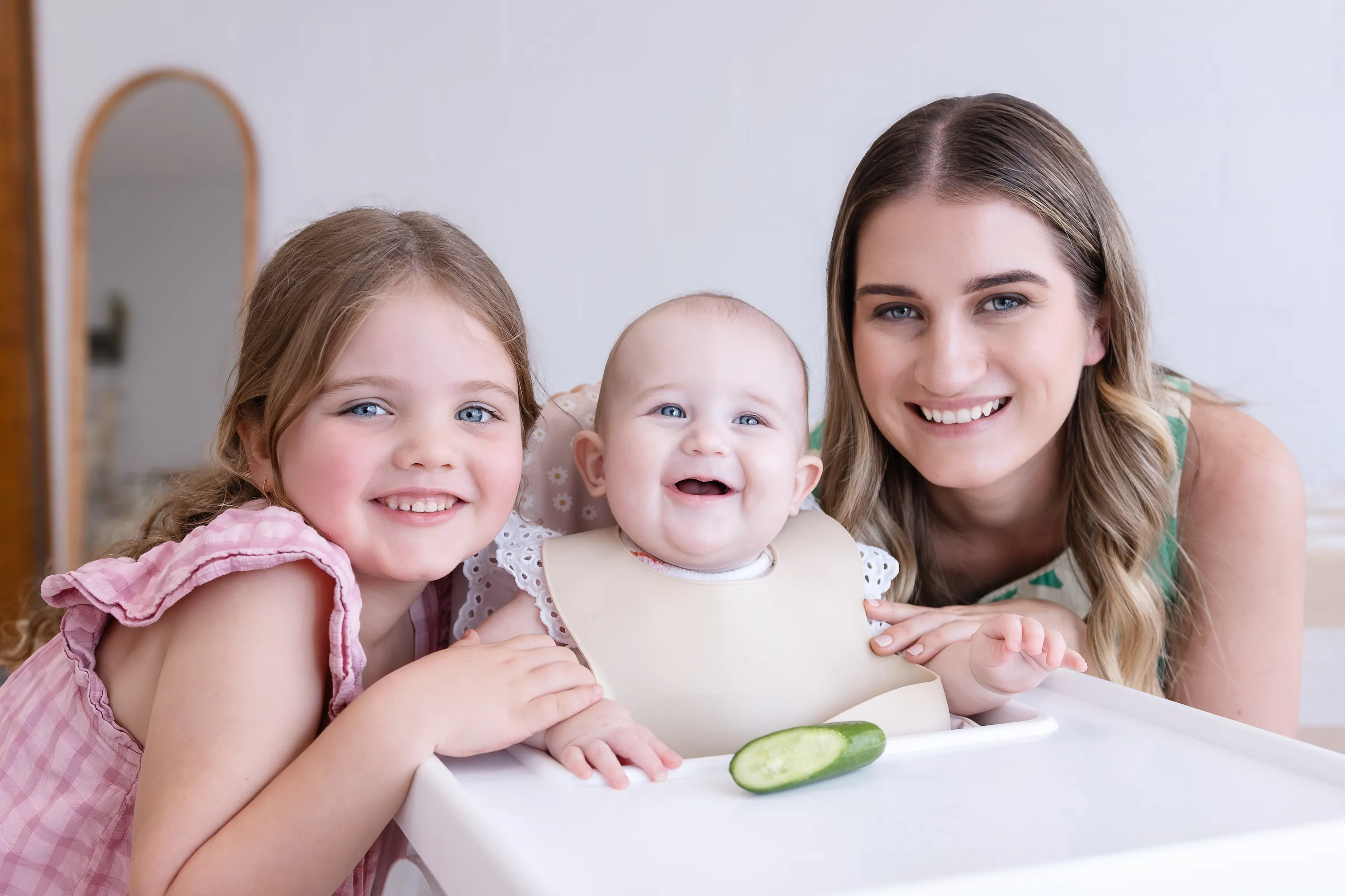 Smiling woman, baby in a high chair with a bib, and a young girl leaning on the high chair with a cucumber slice in front of the baby.