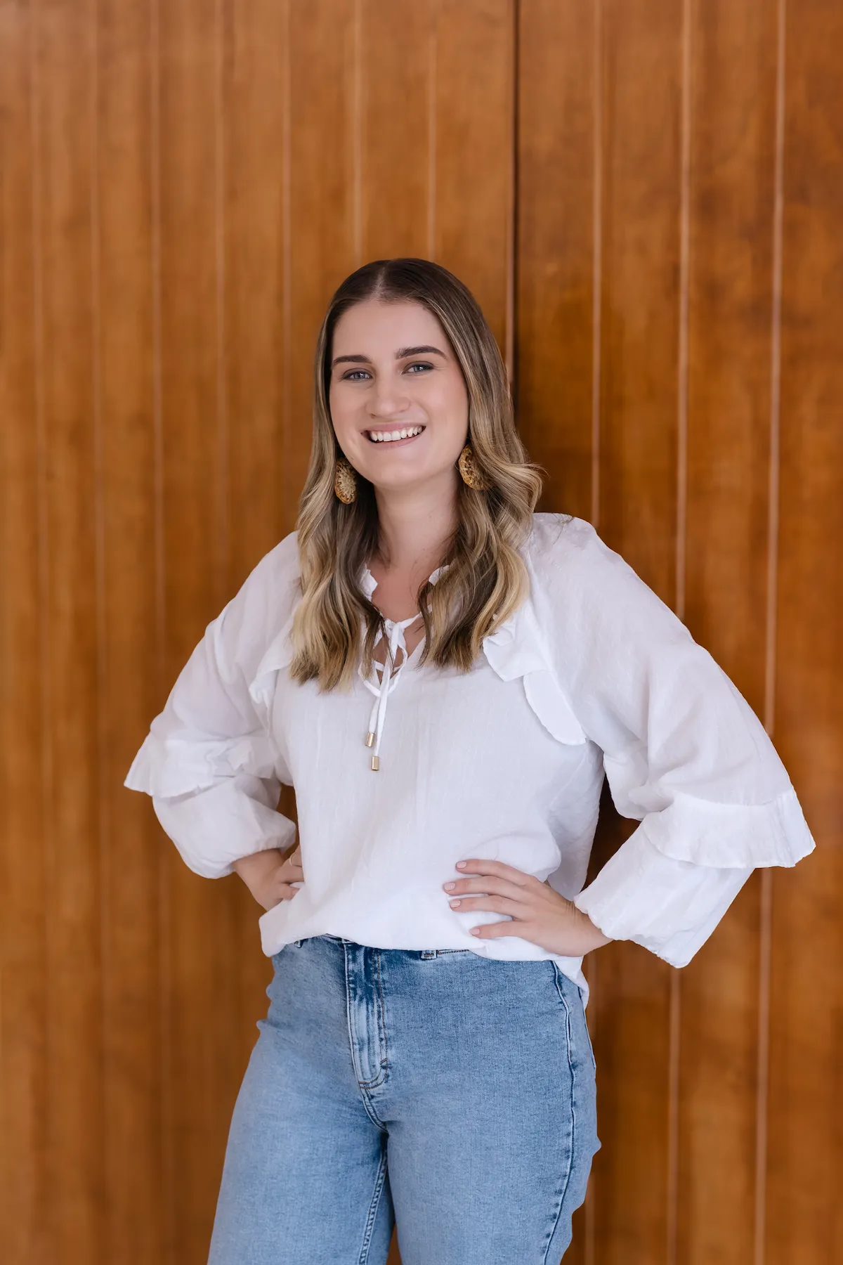 Smiling young woman with wavy blonde hair wearing a white blouse and blue jeans standing with hands on hips against a wooden wall.