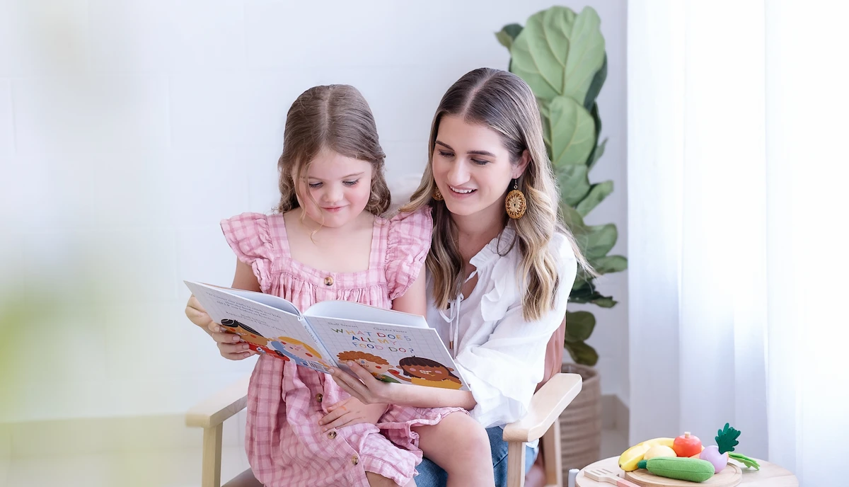 A woman and a young girl sitting on a chair together reading a children's book titled 'What Does All My Food Do?'