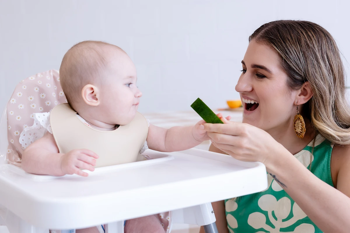 Woman smiling and offering a baby a piece of cucumber while the baby reaches for it in a high chair.