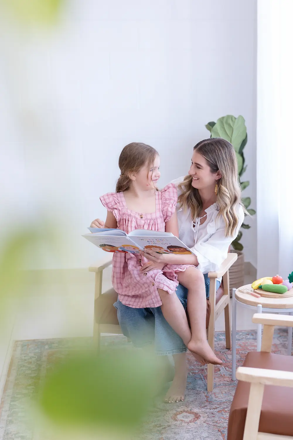 Woman and young girl sitting on a chair together, reading a children's book and smiling at each other in a bright room.