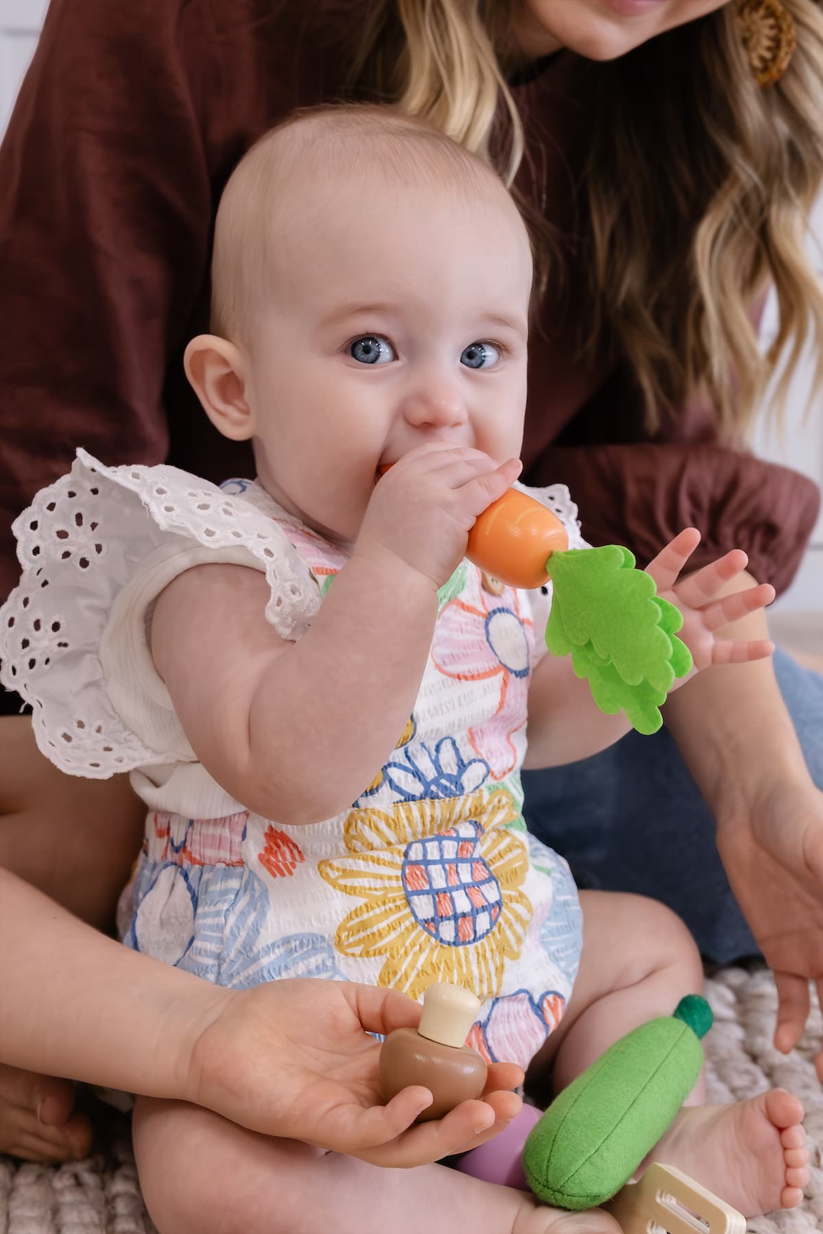 Baby with blue eyes wearing a floral dress, chewing on a toy carrot while sitting on an adult's lap.