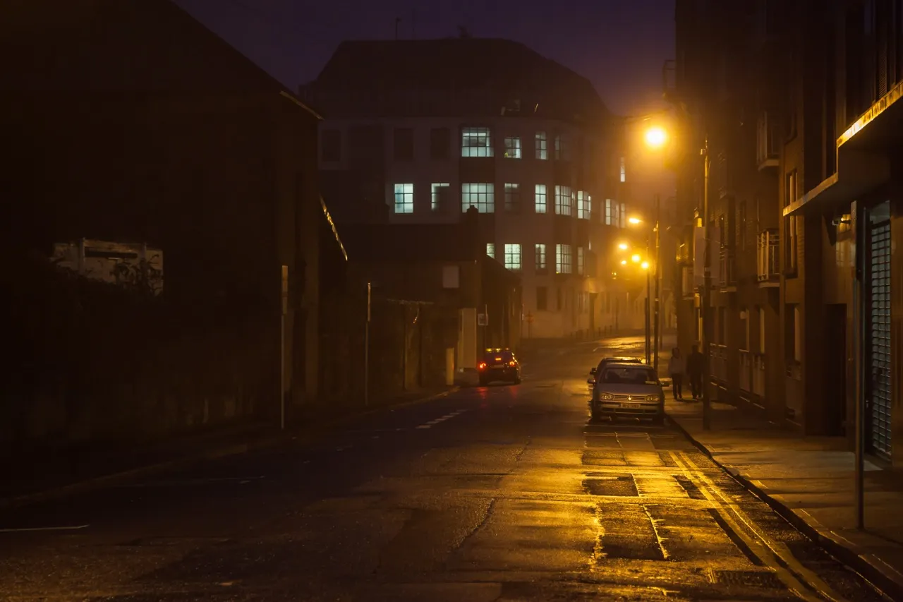 Dimly lit urban street at night with parked cars, illuminated building windows, and yellow streetlights reflecting on wet pavement.