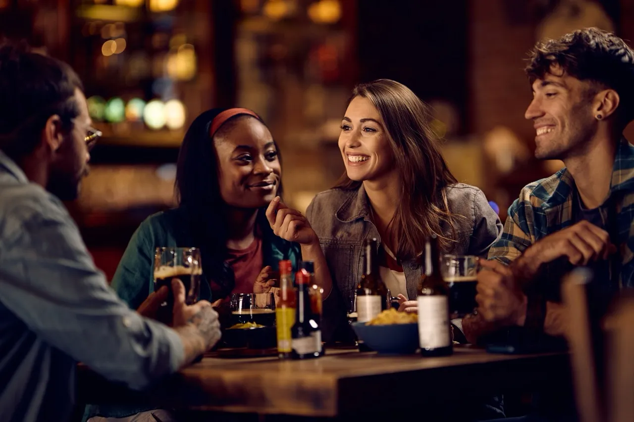 Four friends sitting around a table in a bar, smiling and enjoying drinks together.