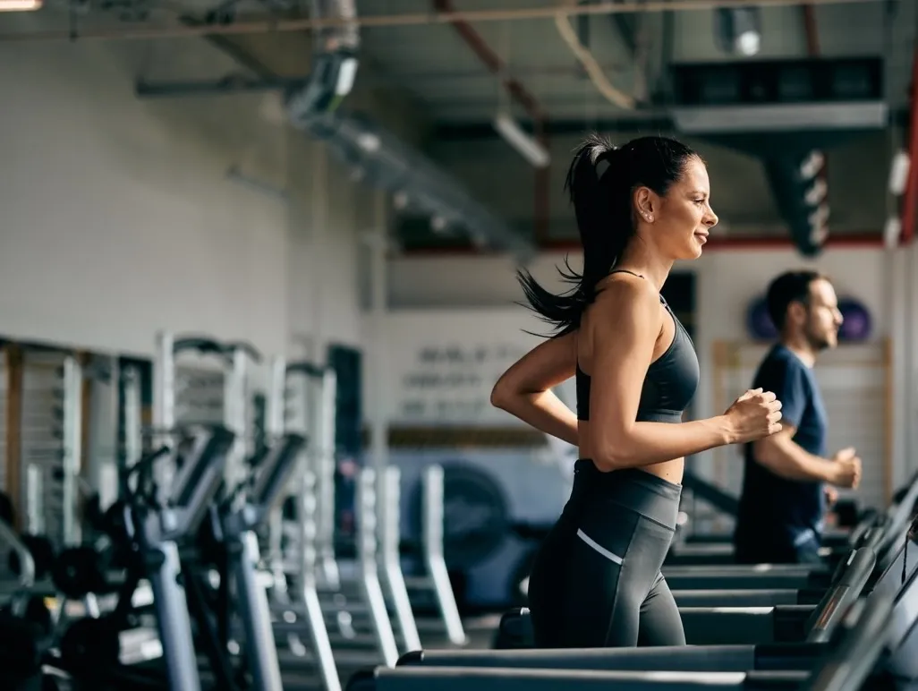 Woman and man jogging on treadmills in a gym with exercise equipment in the background.