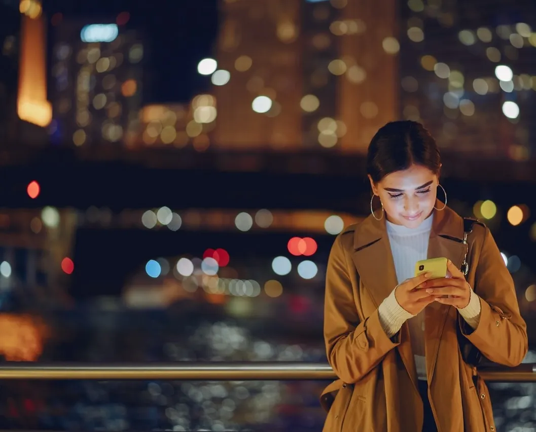 Young woman in a brown coat looking at her phone with city lights blurred in the background at night.