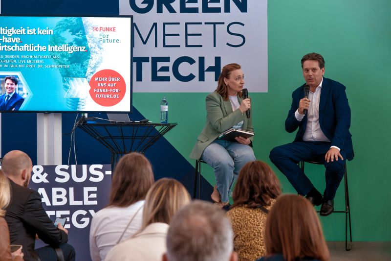 Zwei Personen sitzen auf Barhockern auf einer Bühne, halten Mikrofone und sprechen vor einem Publikum bei einer Veranstaltung mit dem Thema Green Meets Tech.