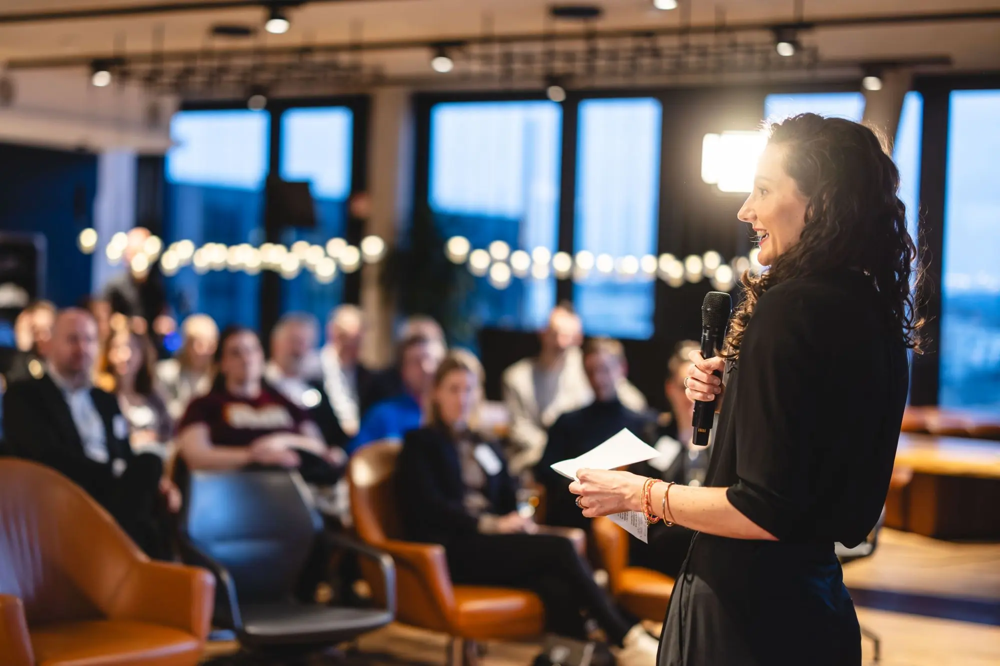 Frau hält Mikrofon und spricht vor einem Publikum in einem modernen Veranstaltungsraum.