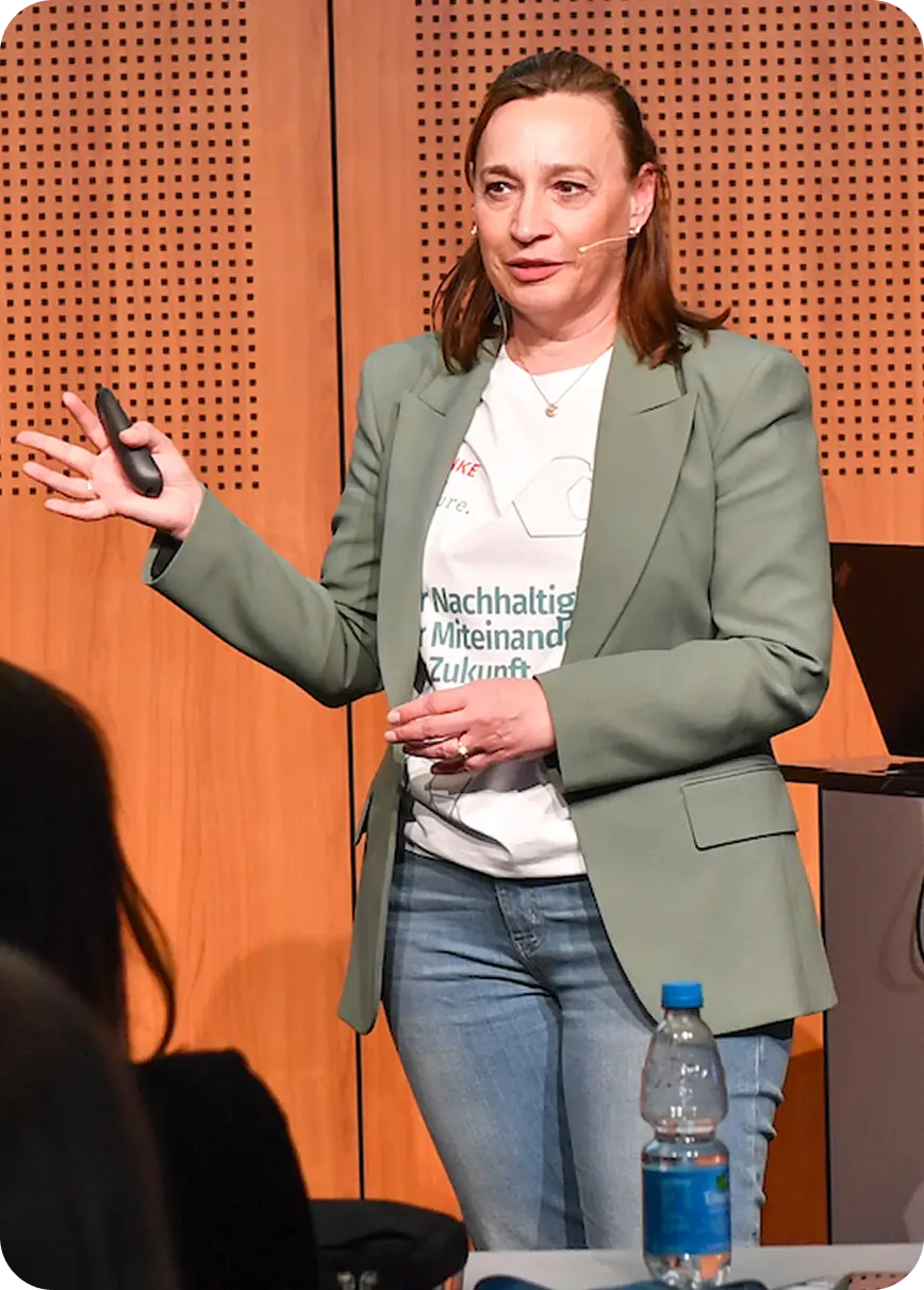 Frau mit Mikrofon am Ohr hält in einem Vortrag eine Präsentation, trägt grünen Blazer und Jeans.