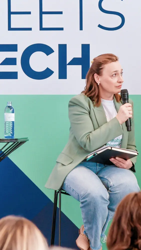 Frau sitzt auf einem Stuhl, spricht in ein Mikrofon und hält ein Notizbuch auf dem Schoß vor einer Wand mit blau-grünem Hintergrund und großen Buchstaben.