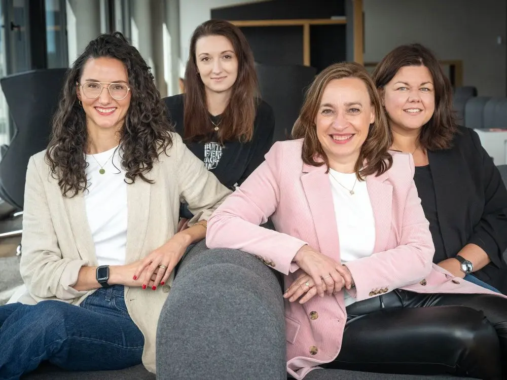 Vier Frauen sitzen auf einem grauen Sofa und lächeln in die Kamera, zwei tragen Blazer.
