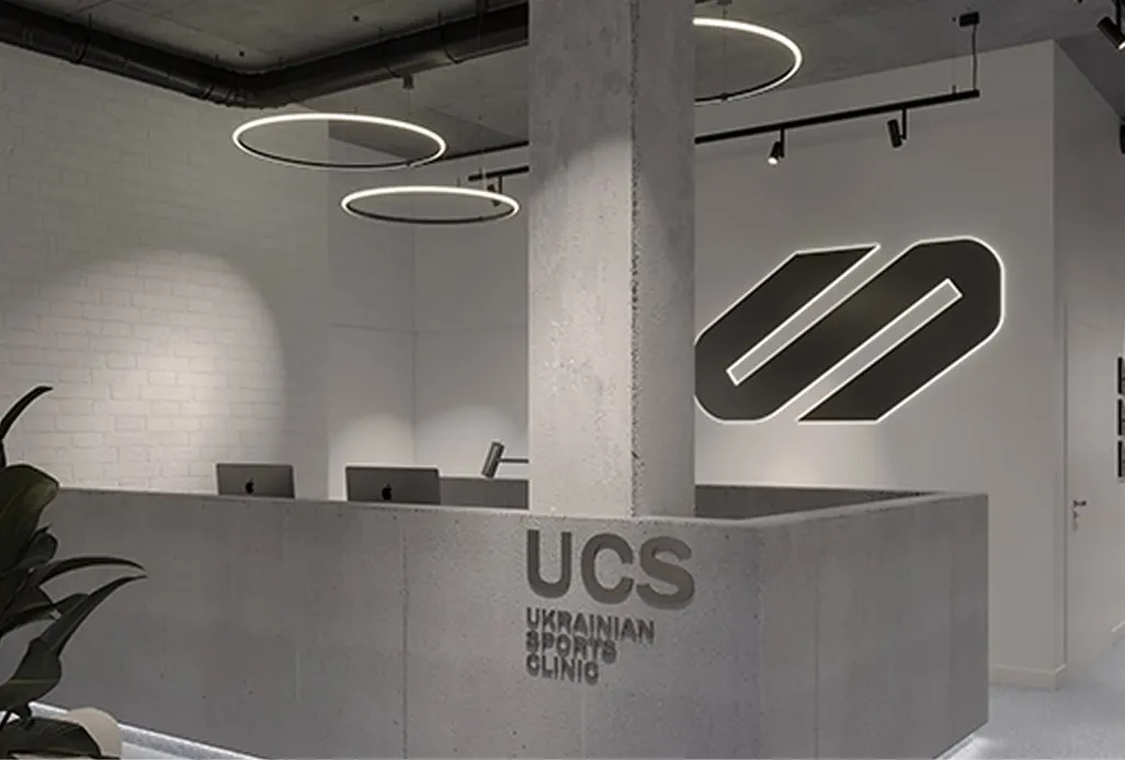 Сучасний мінімалістичний інтер'єр клініки зі стійкою реєстрації, фірмовим логотипом та назвою 'UCS Ukrainian Sports Clinic', ілюструючи консалтинг та бізнес-рішення в спортивній індустрії.