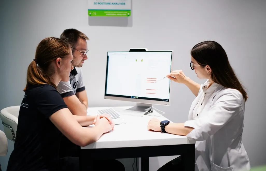 Біомедичний експерт/лікар у білому халаті пояснює результати комплексної діагностики двом клієнтам на комп'ютерному моніторі, підсумовуючи всі дані, зібрані під час чекапів.