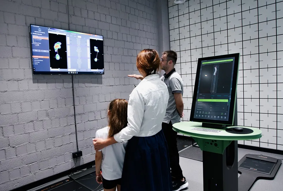 Спеціаліст пояснює сім'ї результати ортопедичного біомеханічного чекапу. На моніторах відображається детальний аналіз постави та розподілу тиску стоп