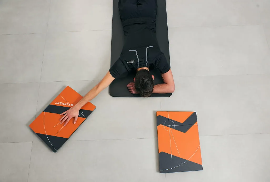 Людина в чорному одязі лежить обличчям донизу на сірому килимку, тягнучись до оранжево-чорної книги на кахляній підлозі. Сцена передає стан зосередженості.