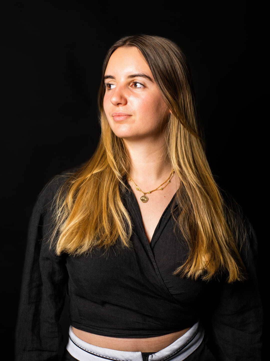 Jeune femme avec cheveux longs blonds portant un haut noir et un collier doré, regardant vers la gauche sur fond noir.
