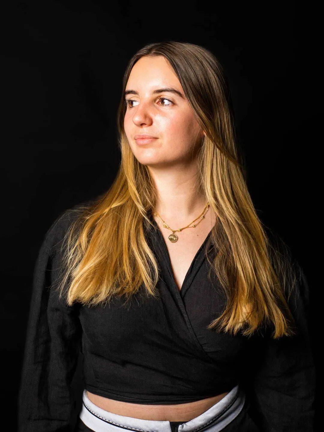 Jeune femme avec cheveux longs blonds portant un haut noir et un collier doré, regardant vers la gauche sur fond noir.