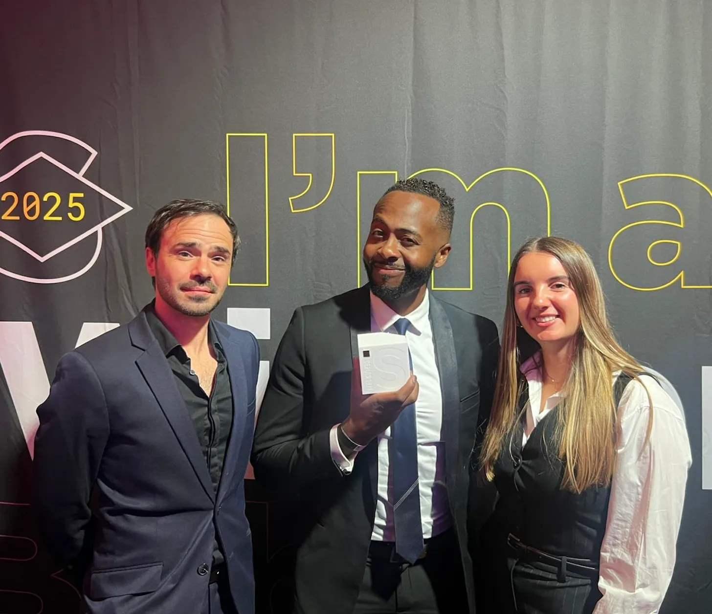 Trois personnes souriantes posant devant une bannière noire, un homme au centre tient un trophée blanc.