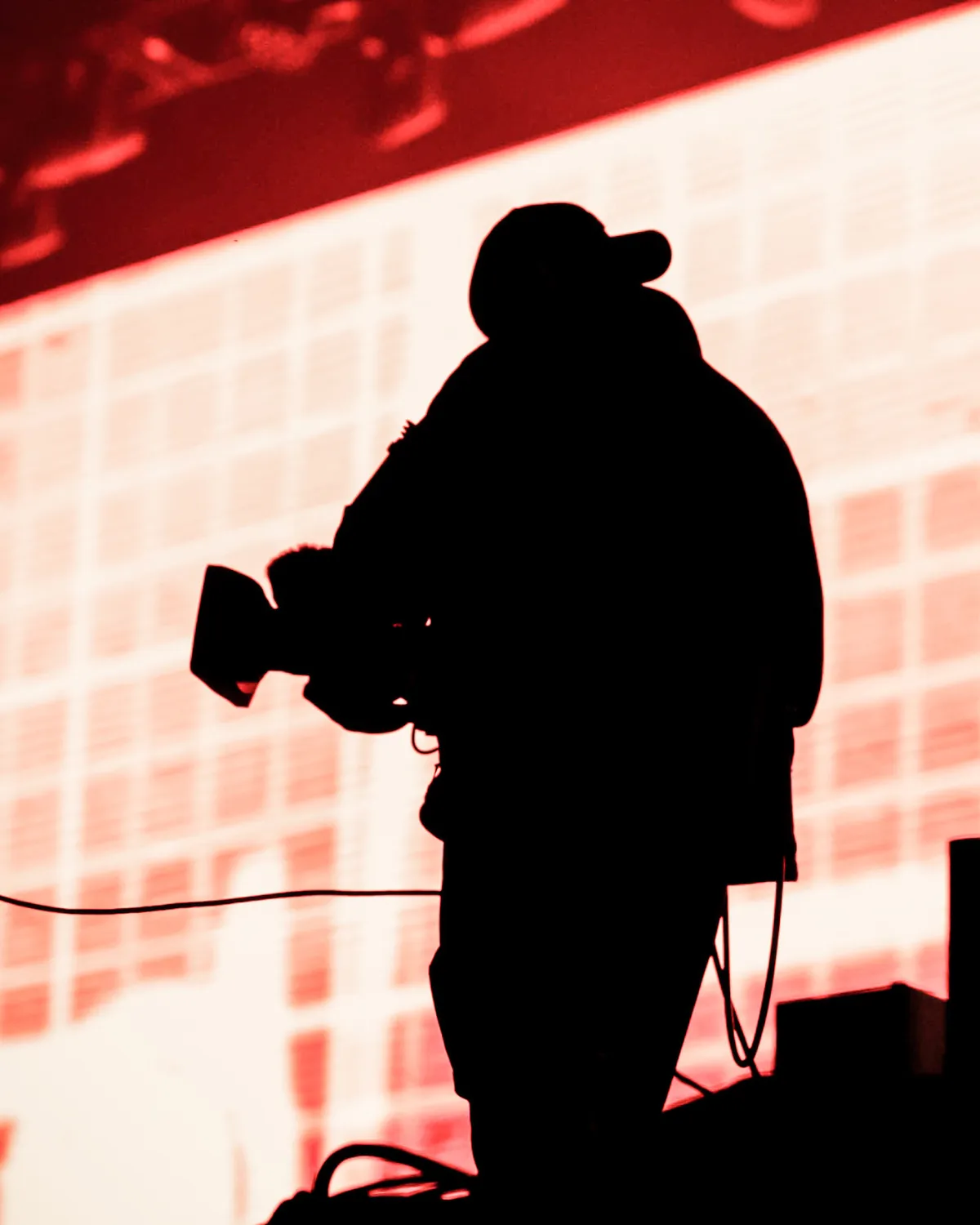 Silhouette d'un caméraman portant une casquette, tenant une caméra vidéo contre un fond lumineux rouge.