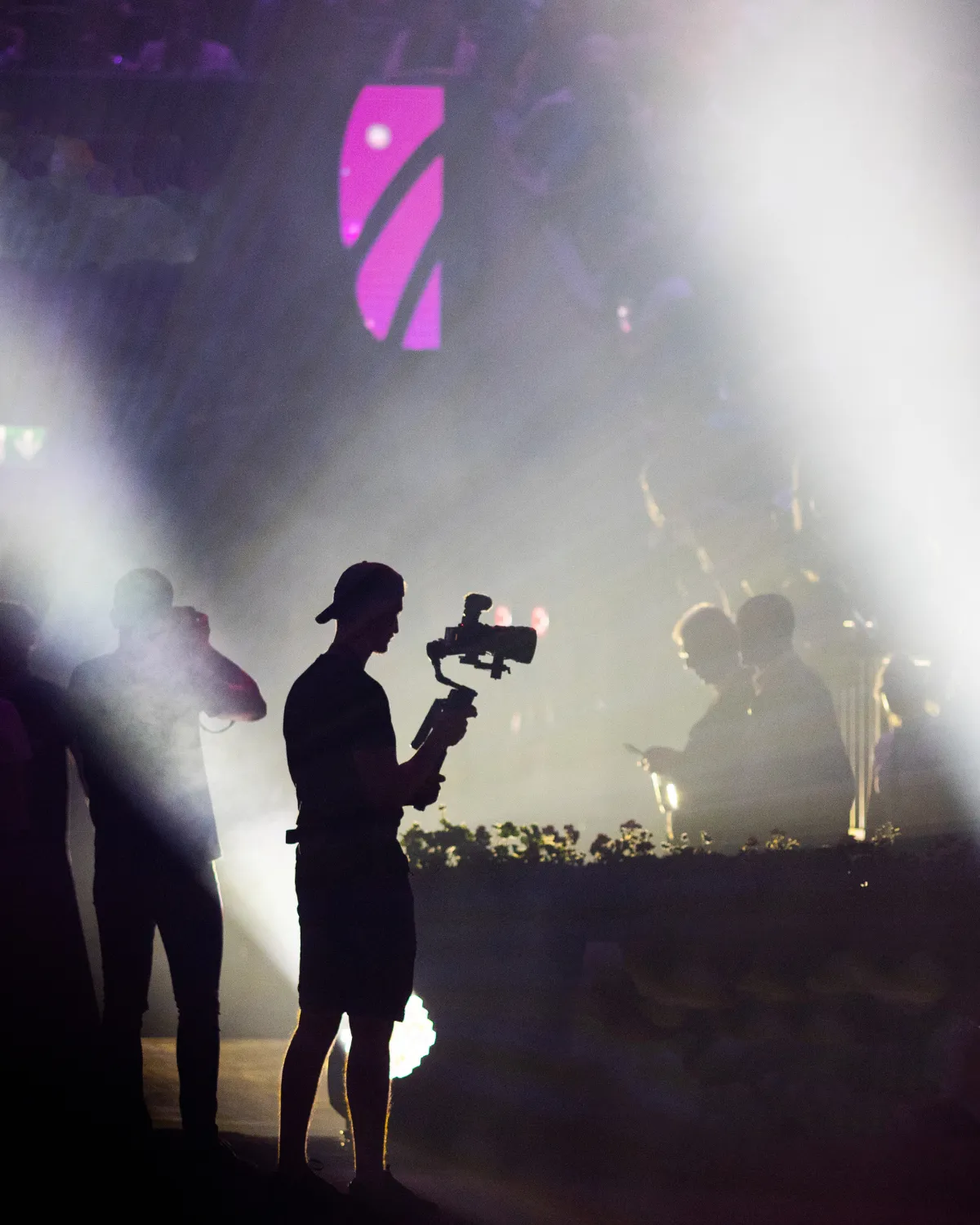 Silhouette d'un caméraman tenant une caméra stabilisée lors d'un événement avec des faisceaux lumineux en arrière-plan.