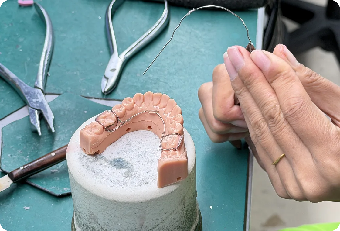 Hands shaping a wire component beside a dental mold of upper teeth with dental tools on a work surface.