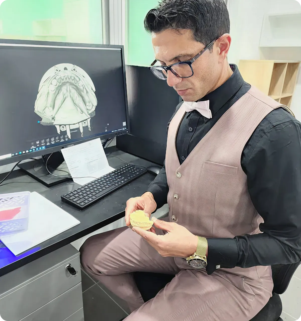 Dr. Sanad Al Murayati in glasses and bowtie examining a dental mold while a computer screen displays a 3D dental model.